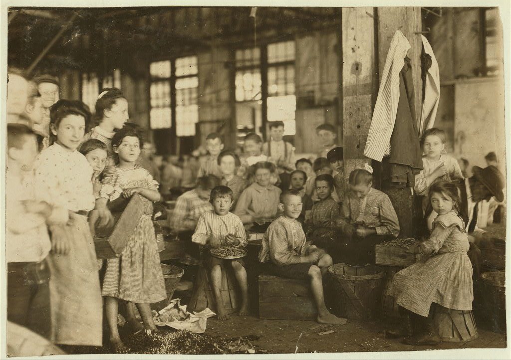 Children and women sit on crates and baskets stringing beans inside a cannery shed.